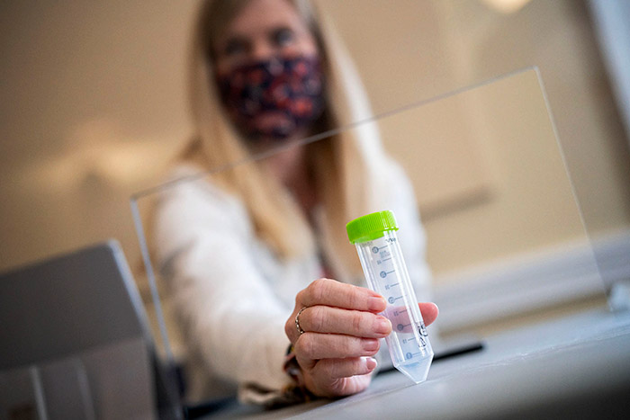 A masked lab technician holding a plastic vial close to the foreground