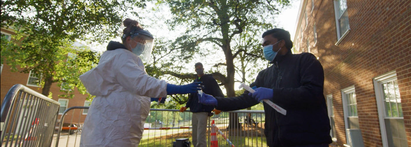 An employee in full-coverage PPE handing a sampling vial to a scientist wearing latex gloves and a face mask