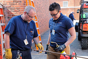 A man in yellow work gloves and a woman holding an unlit torch examine a copper pipe